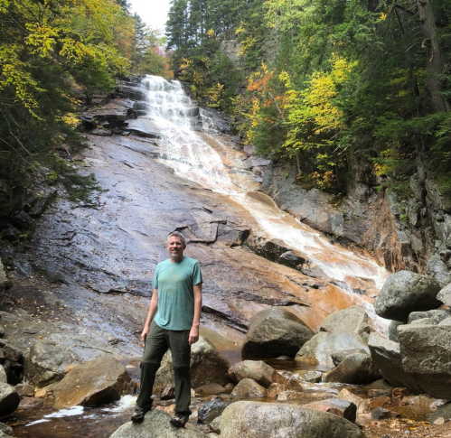 A person stands on rocks in front of a waterfall surrounded by trees with autumn foliage.