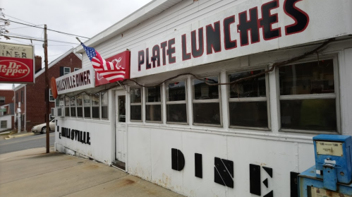 Exterior of a diner with a flag, featuring signs for plate lunches and a vintage blue vending machine.