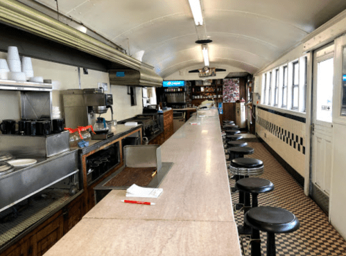 Interior of a vintage diner with a long counter, stools, and a kitchen area, featuring checkered flooring and bright lighting.