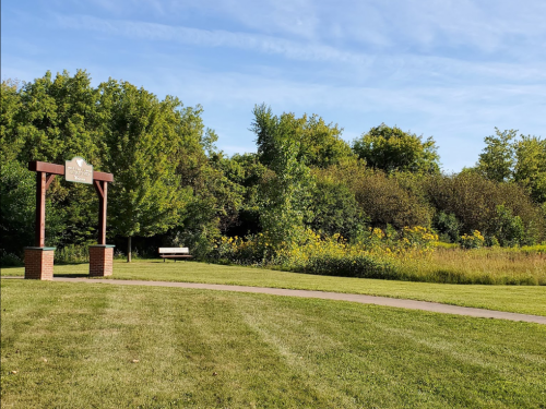 A park scene featuring a sign, a bench, and lush greenery under a clear blue sky.