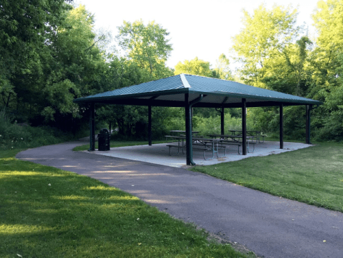 A shaded picnic shelter with tables, surrounded by trees and a paved path in a park setting.