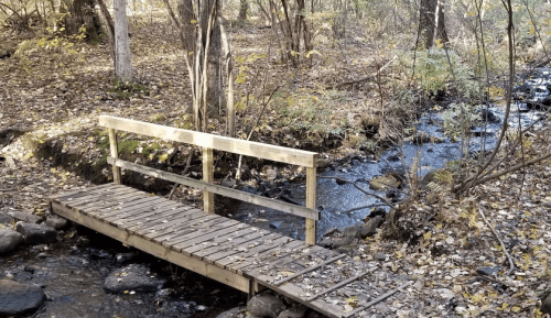 A wooden bridge over a small stream, surrounded by trees and autumn leaves.