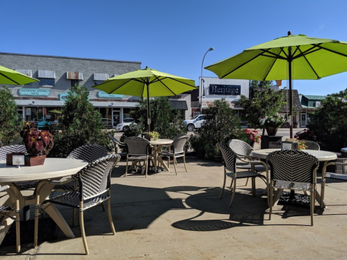 Outdoor café scene with tables and green umbrellas, surrounded by plants and flowers, under a clear blue sky.