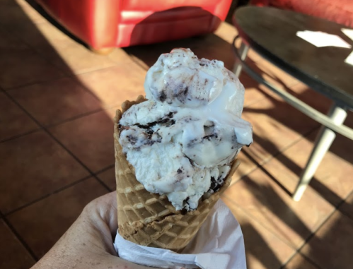 A hand holding a waffle cone filled with swirled chocolate and vanilla ice cream, with a cozy café background.