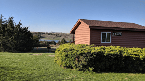 A brown cabin labeled "4 Goose" surrounded by greenery, with a lake visible in the background under a clear blue sky.