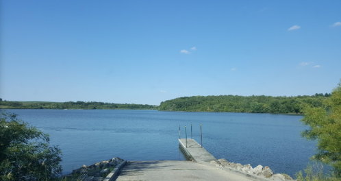 A serene lake view with a wooden dock extending into the water, surrounded by greenery and a clear blue sky.