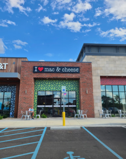 Exterior of a restaurant with a sign reading "I ❤️ mac & cheese," featuring outdoor seating and a blue sky.