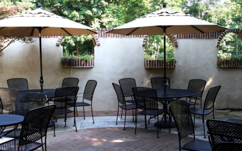 Outdoor dining area with black metal tables and chairs, shaded by large umbrellas, surrounded by greenery and brick walls.