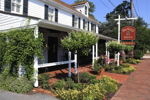 A charming white restaurant with green shutters, surrounded by colorful flowers and plants, featuring a sign for Buckley's Tavern.