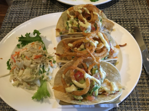 Three tacos topped with avocado, tomatoes, and crispy onions, served with a side of coleslaw on a white plate.