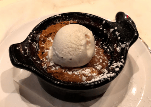 A warm dessert in a black dish topped with a scoop of vanilla ice cream and a dusting of powdered sugar.