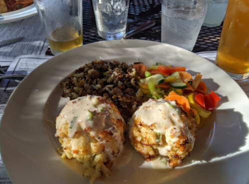 A plate featuring two crab cakes with sauce, a side of wild rice, and a colorful vegetable medley.