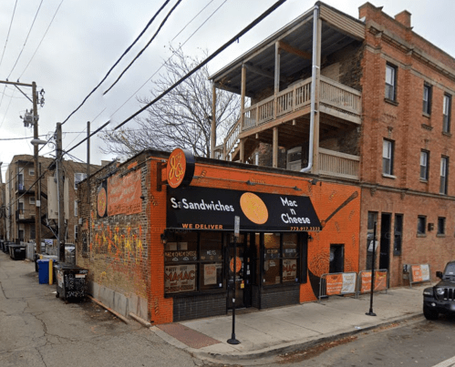 A corner restaurant with a bright orange sign reading "Mac n Cheese" and outdoor seating, surrounded by brick buildings.