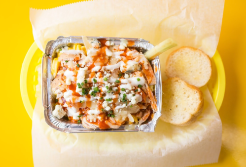 A foil tray filled with colorful toppings, drizzled sauce, and two slices of bread on a yellow background.