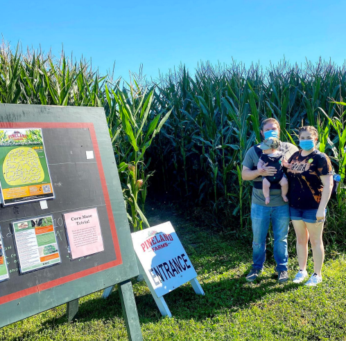 A family stands by a corn maze entrance sign, wearing masks, with tall corn plants in the background under a clear blue sky.
