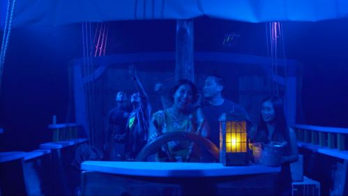 A group of people on a boat set, with one person at the helm, illuminated by blue lighting and a lantern.