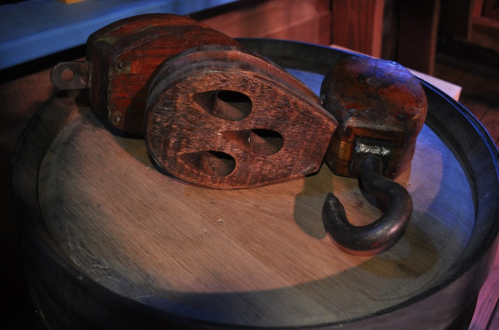 A wooden pulley with a hook resting on a wooden barrel, illuminated with soft lighting.