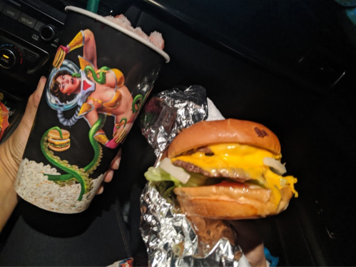 A hand holds a colorful drink cup featuring a graphic design, alongside a cheeseburger wrapped in foil.