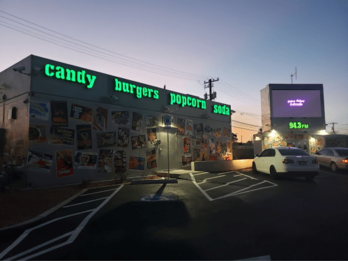 A colorful building with signs for candy, burgers, popcorn, and soda, surrounded by vintage movie posters at dusk.