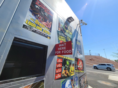 A food truck with vintage horror movie posters and a sign that says "WAIT HERE FOR FOOD." Sunny outdoor setting.