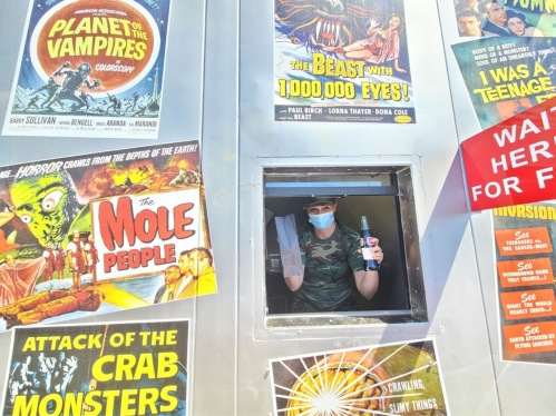 A person in a mask holds a drink at a window surrounded by vintage horror movie posters.