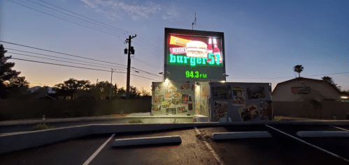 A brightly lit burger restaurant sign at dusk, featuring a large burger graphic and the radio station 94.3 FM.