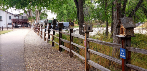 A pathway lined with birdhouses on a wooden fence, surrounded by trees and greenery.