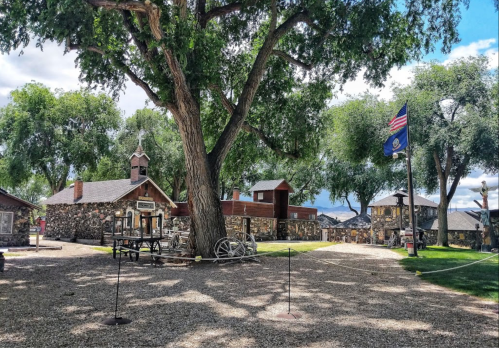Historic site with a large tree, stone buildings, and an American flag under a blue sky with scattered clouds.