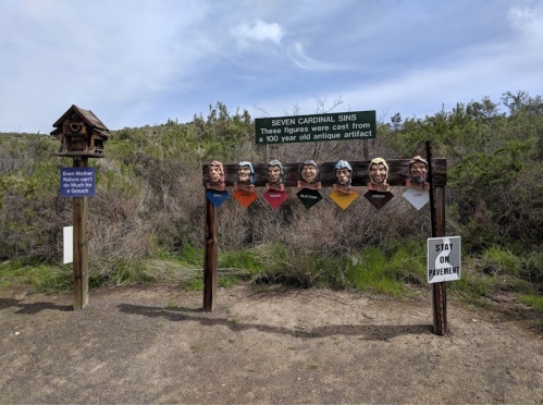 Sign displaying the "Seven Cardinal Sins" with carved figures, set in a natural landscape with a birdhouse nearby.
