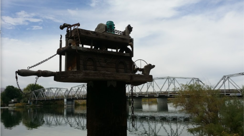 A whimsical wooden sculpture of a truck on a post, overlooking a calm river with a bridge in the background.