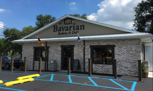 Exterior of Bavarian Bakery & Deli, featuring a brick facade, flags, and a blue parking lot with yellow barriers.