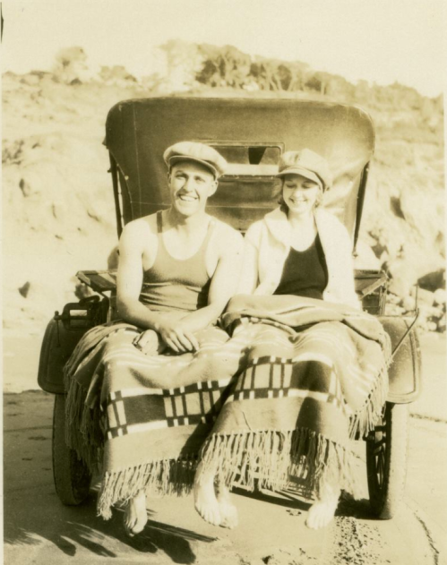 A vintage photo of a man and woman in swimwear, sitting on a blanket in an open car by the beach.