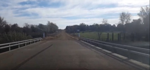 A view of a rural road leading to a bridge, surrounded by trees and open fields under a cloudy sky.