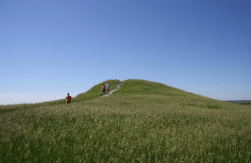 Two people walk up a grassy hill under a clear blue sky, with steps leading to the top.
