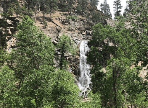 A waterfall cascades down rocky cliffs, surrounded by lush green trees and rugged terrain.