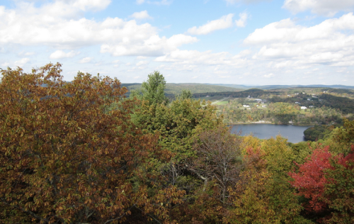 A scenic view of rolling hills and a lake, surrounded by trees displaying autumn colors under a partly cloudy sky.