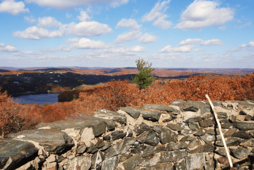 A scenic view of autumn foliage and a river, with a stone wall in the foreground and blue skies above.