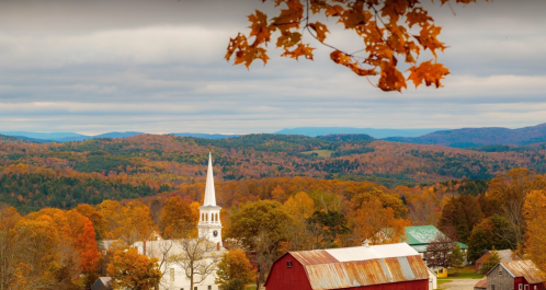 A scenic view of a church steeple surrounded by vibrant autumn foliage and rolling hills under a cloudy sky.