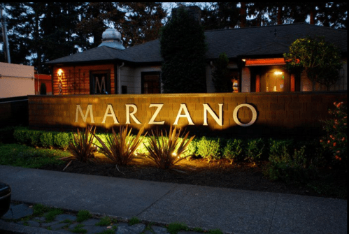 Illuminated sign for Marzano restaurant, surrounded by greenery and a cozy building in the background.