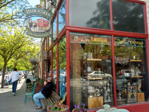 A storefront with a sign reading "Hyde & Seek" featuring unique goods, surrounded by trees and pedestrians.