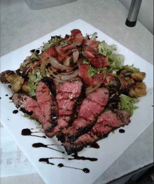 A plate of sliced steak on a bed of greens, topped with bacon, grilled onions, and drizzled with balsamic glaze.