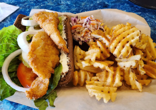 A fried shrimp sandwich with lettuce, tomato, and onions, served with coleslaw and waffle fries on the side.