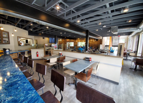 Modern restaurant interior with a blue marble counter, wooden accents, and empty tables under industrial-style lighting.