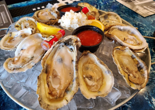 A platter of fresh oysters on ice, garnished with lemon wedges, cocktail sauce, and hot sauce.