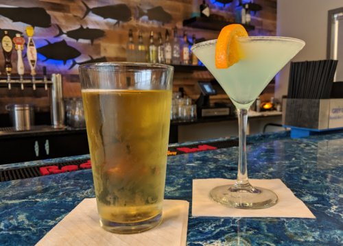 A glass of beer and a blue cocktail with an orange slice sit on a bar counter with a wooden backdrop.