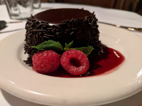A chocolate dessert topped with chocolate sprinkles, garnished with fresh raspberries and mint on a white plate.