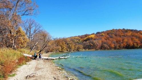 A serene lakeside scene with two people walking along a sandy shore, surrounded by colorful autumn foliage.
