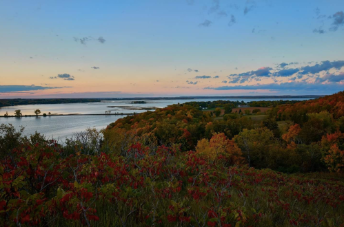 A scenic view of a river surrounded by vibrant autumn foliage under a colorful sunset sky.