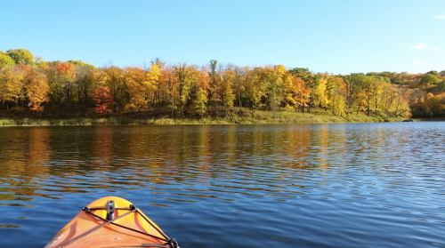 A kayak on calm water reflects vibrant autumn trees in shades of orange, yellow, and green under a clear blue sky.