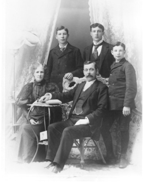 A vintage black-and-white photo of a family: a seated man with a book, flanked by three young boys and a woman.